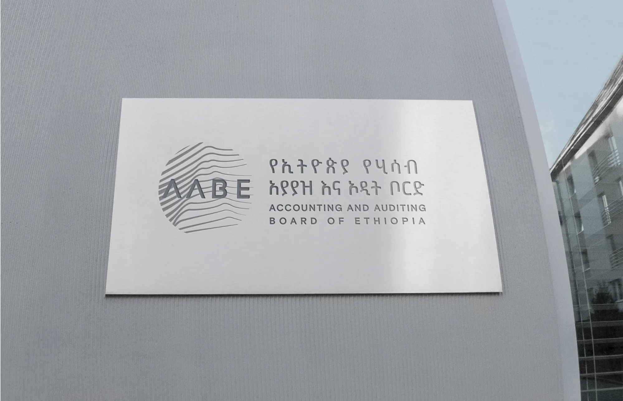A brushed-metal plaque on a grey wall, with the bilingual AABE logo engraved into it, beside a glass building.