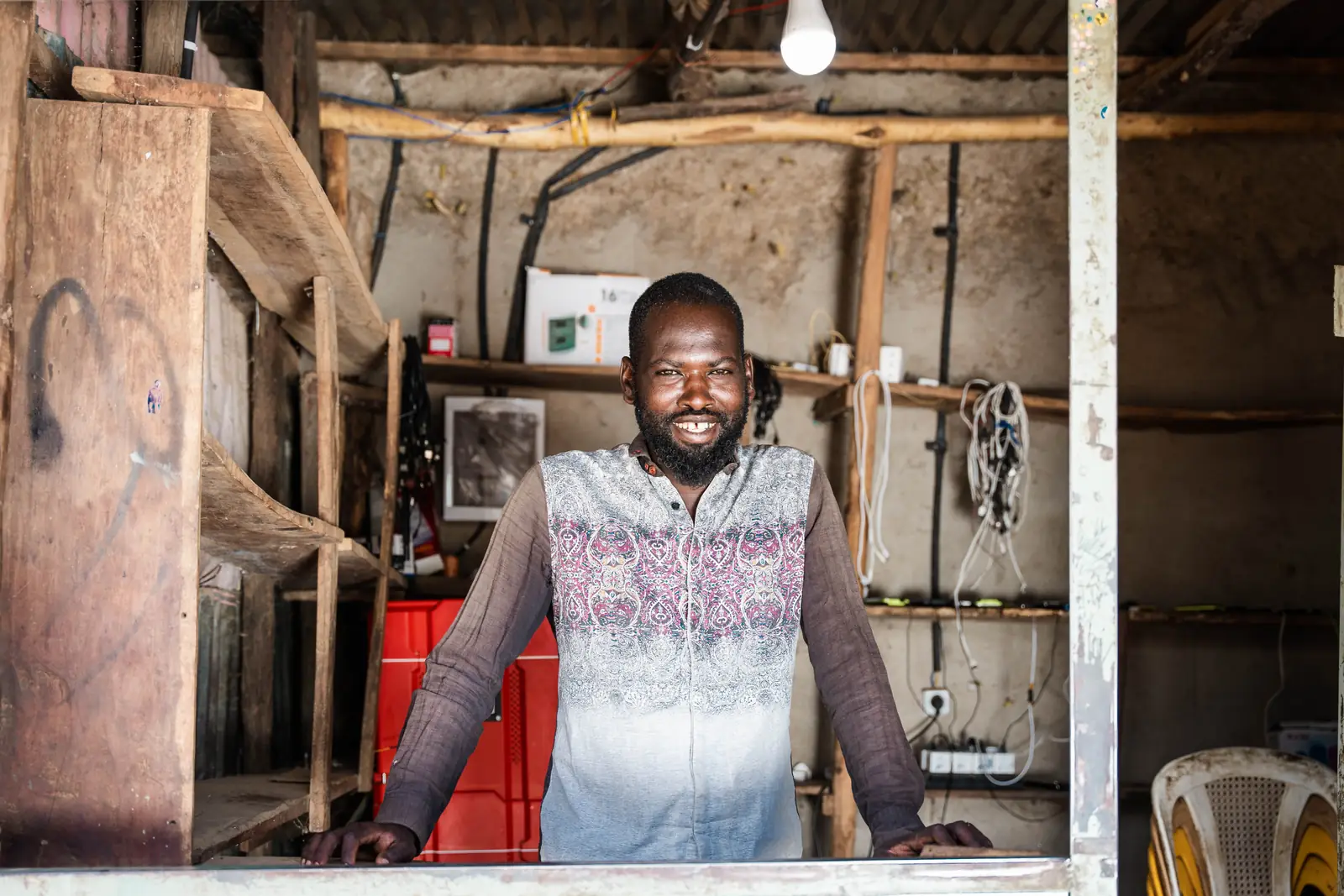 Mr. Amanuel stands behind the counter of his wooden shop, shelves of equipment and charging cables behind him.