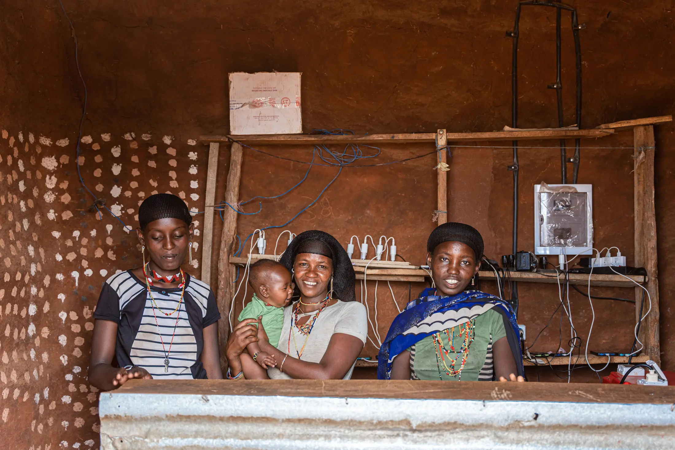 Three women and a baby behind the counter of a mud-walled charging kiosk in Borana, phones charging on the shelf behind them and a Powerblox gateway box on the wall.
