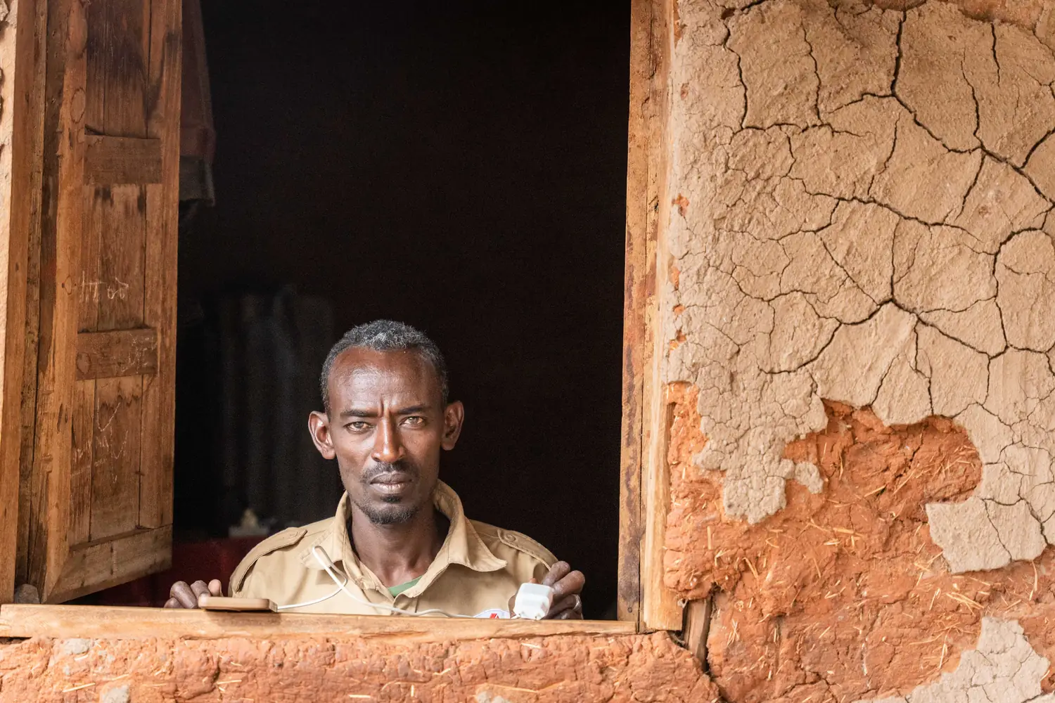 A man looks out through the wooden window of a cracked mud-plaster building.
