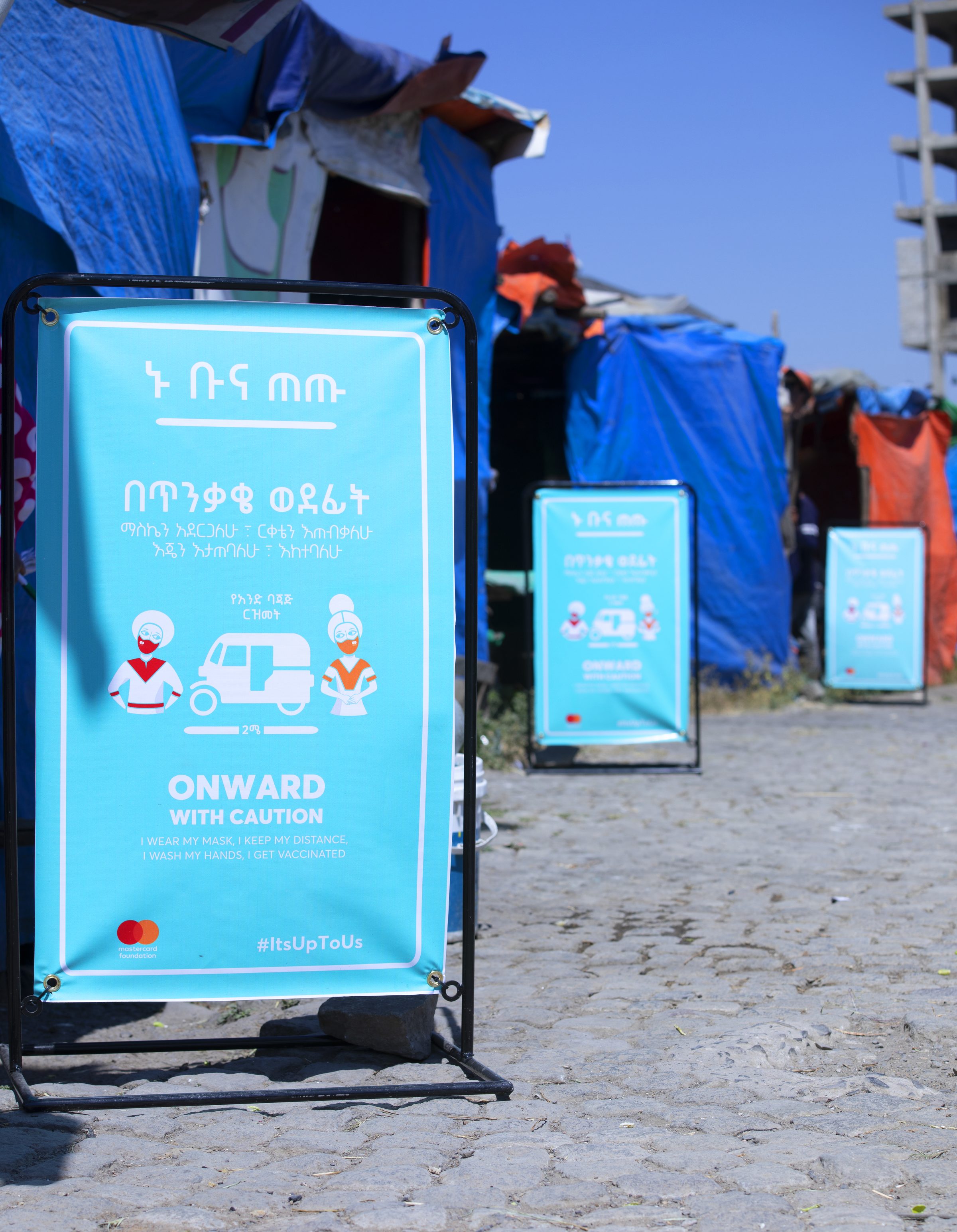 A turquoise Onward with Caution street banner on a stand beside coffee stalls, with masked figures and a three-wheeled taxi illustrating physical distance.