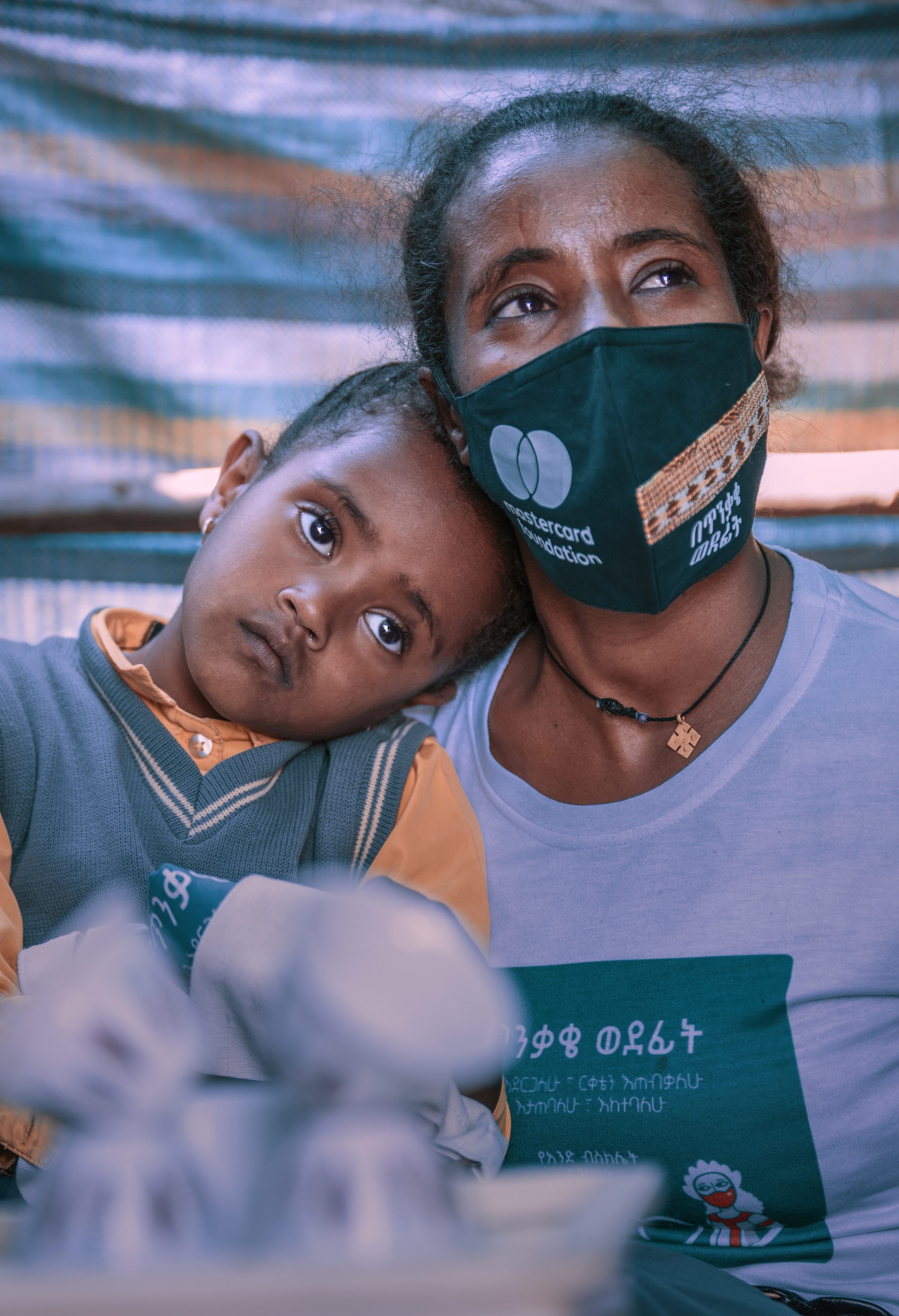 A coffee vendor in a Mastercard Foundation face mask and campaign T-shirt, her young daughter resting her head on her shoulder.
