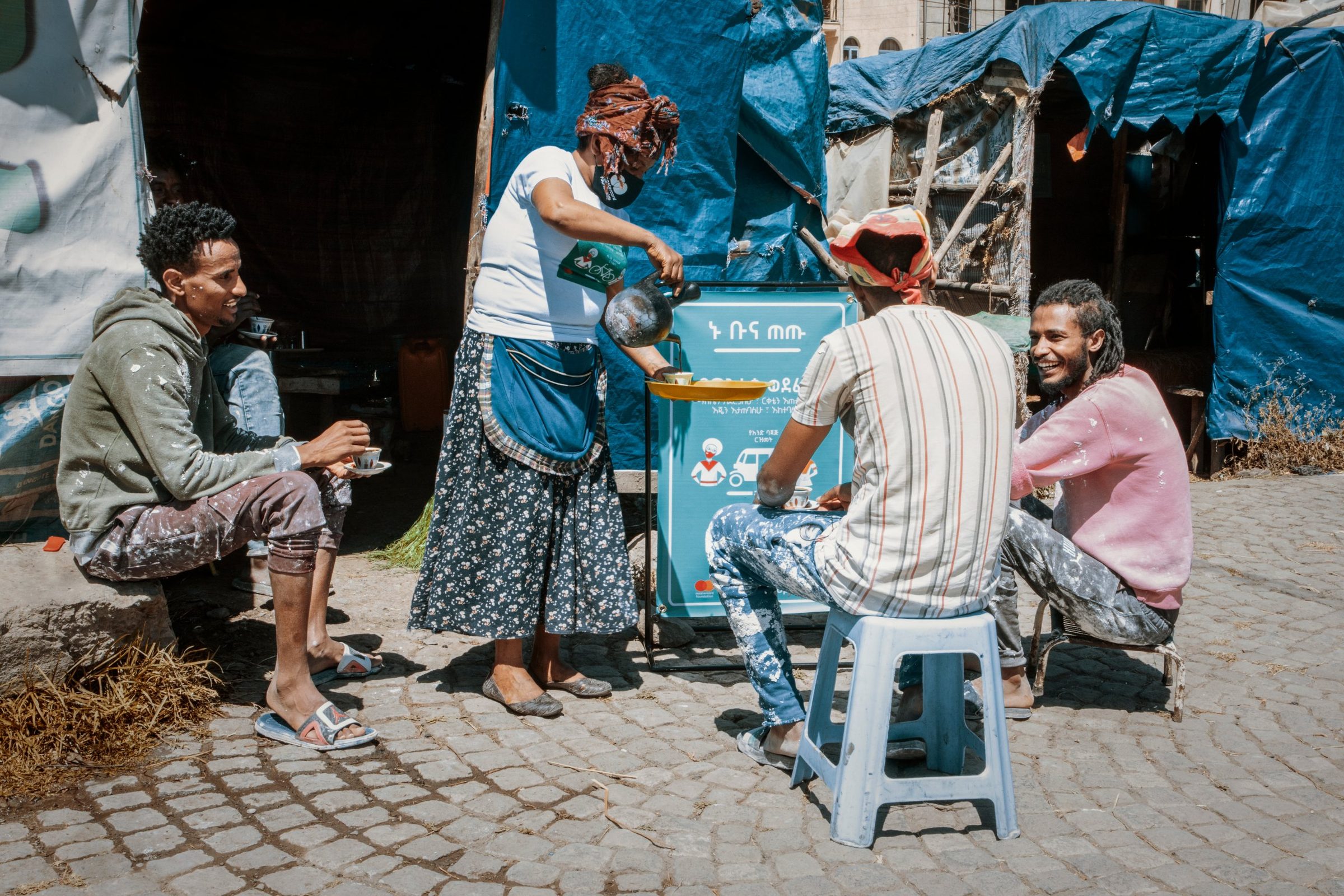 A coffee vendor pours coffee for customers at her street stall, a turquoise campaign banner beside her.