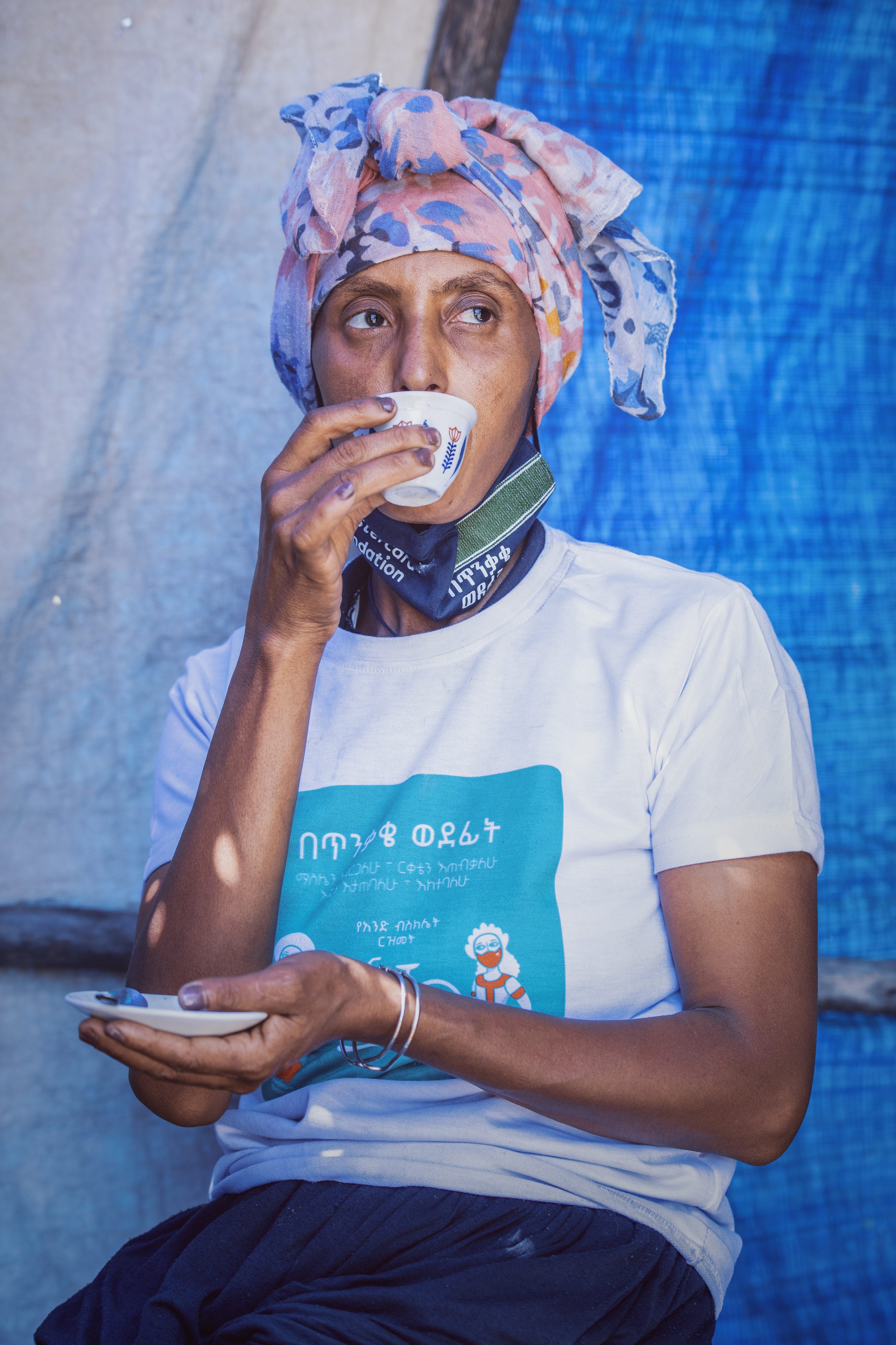 A coffee vendor in a patterned headwrap and campaign T-shirt drinking from a small coffee cup.