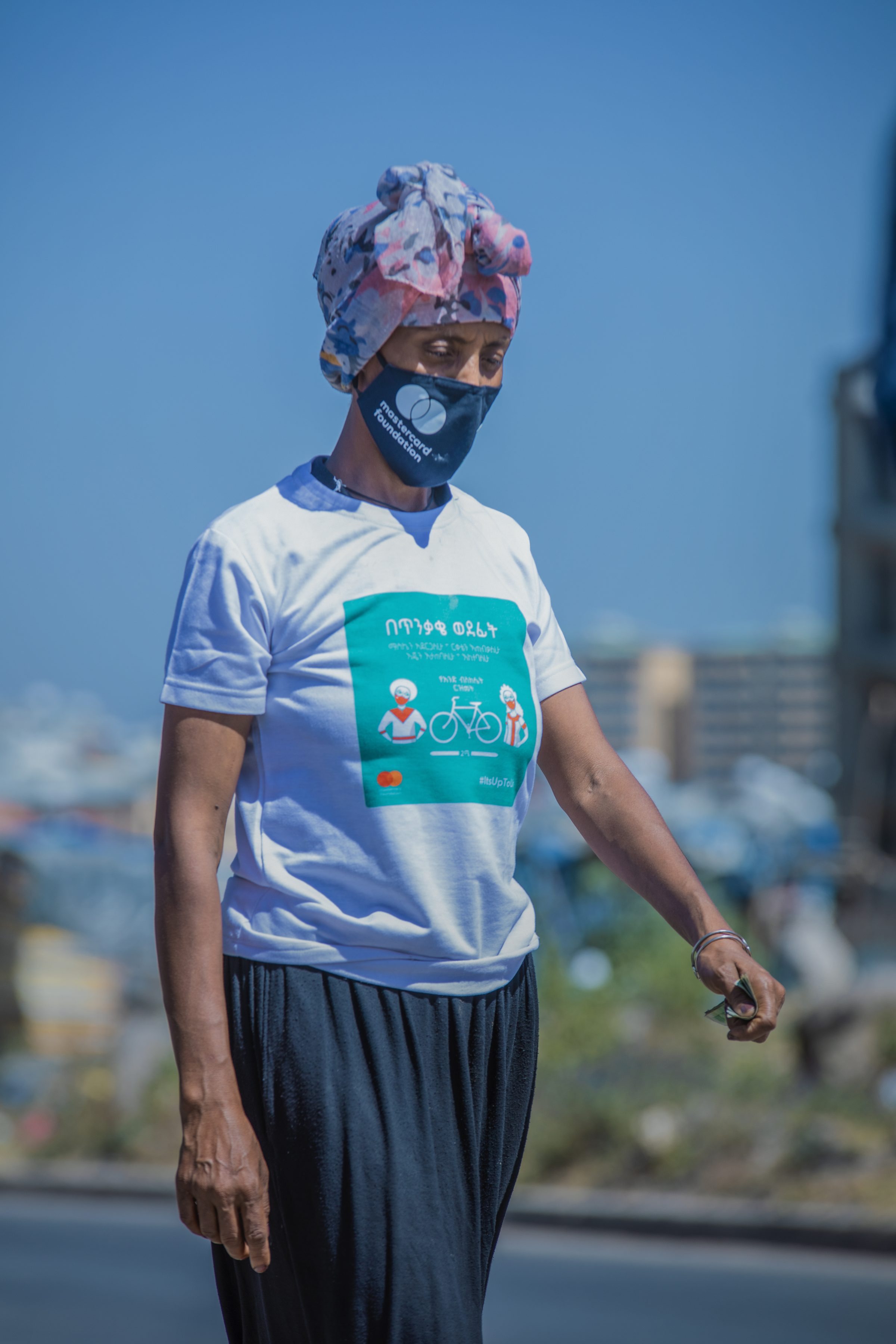 A coffee vendor in a campaign T-shirt and mask walks along a street.
