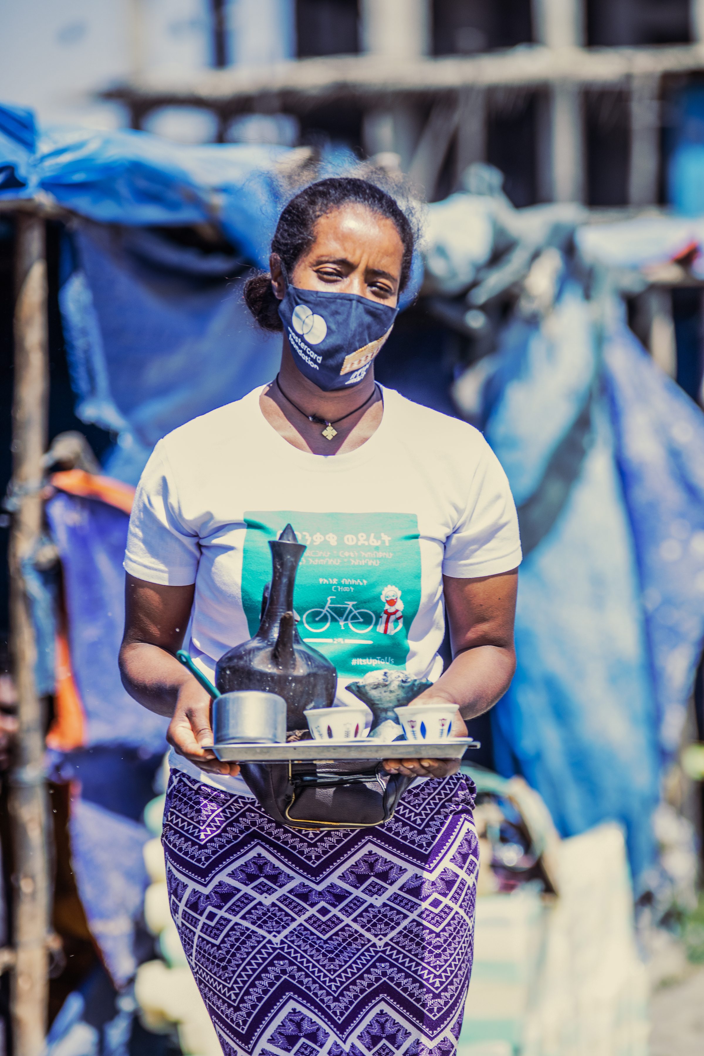 A coffee vendor in campaign T-shirt and mask holds a tray of coffee cups.